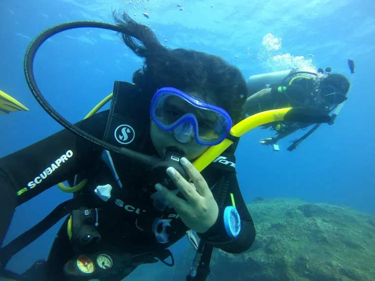 Two divers underwater in blue water in wetsuits, masks and tanks, one reaching towards the camera.