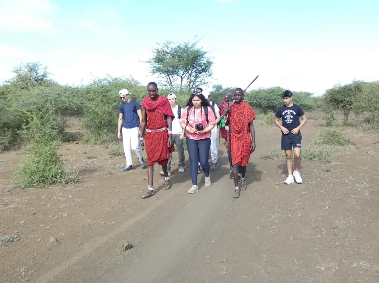 Shonairra and classmates walking a dirt road in Kenya with two Maasai guides in red shukas, thorn scrub on either side.